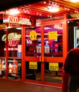 A brightly lit entrance to a casino with neon signs, including one advertising a £500 jackpot. The doors have clear signage indicating entry is restricted to those over 18. A person in a red jacket stands to the right, possibly interacting with a machine or display.